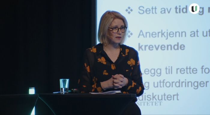 Professor Anniken Furberg står på en scene med en PowerPoint-presentasjon bak seg.
