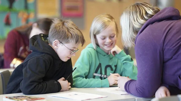 to barn som sitter og får hjelp av en lærer i et klasserom, de smiler. 