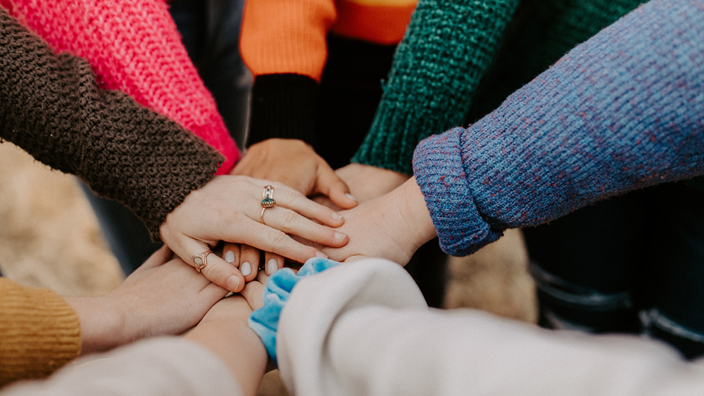 Nærbilde: av en gruppe ulike mennesker holder hender og er kledd i gensere i mange ulike farger. Illustrasjonsfoto.
