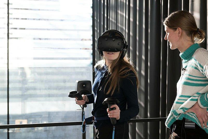 Two persons in a room, one wears a virtual reality headset and holds two touch controllers.