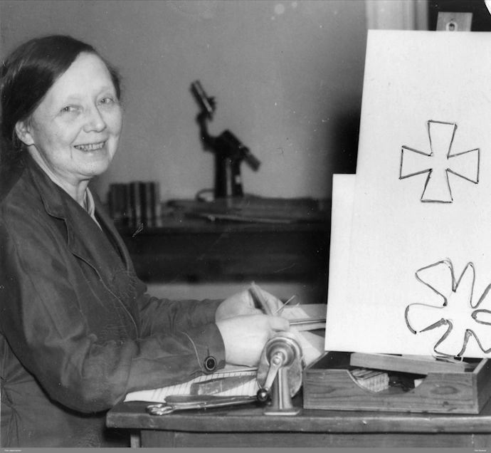 Eldre fotografi av kvinnen Helga Eng sittende ved skrivebord. Svart-hvitt. Barnetegninger i front.
