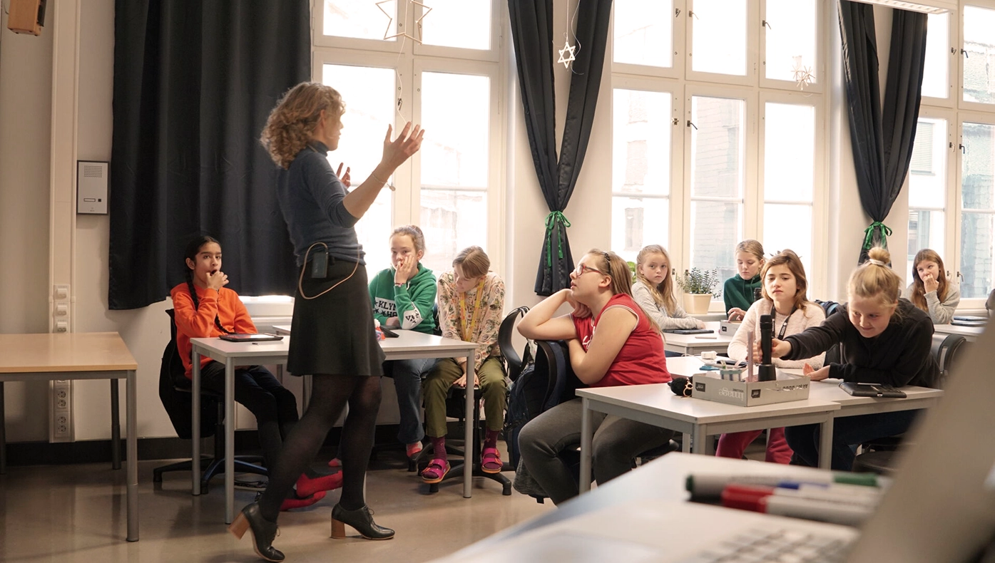 Kvinnelig lærer står foran klasse i klasserom og gestikulerer.