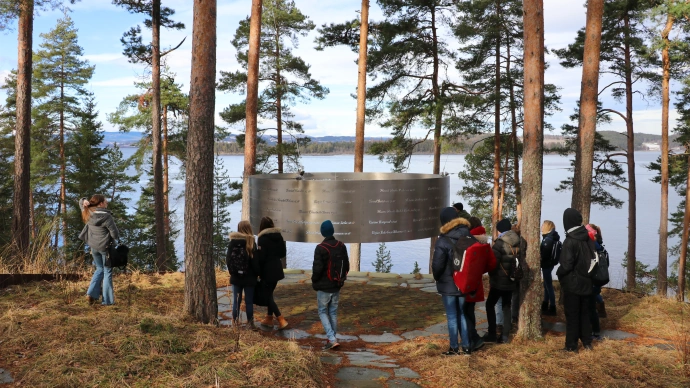Ungdom står kring eit av minnesmerka på Utøya; ein stor ring som heng mellom trea og har namna på dei som vart drepne 22. juli skrive på seg.