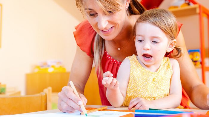 Bilde av barnehagelærer og et barn som sitter sammen. Barnehagelæreren tegner med en grønn blyant mens barnet ser på og virker engasjert.