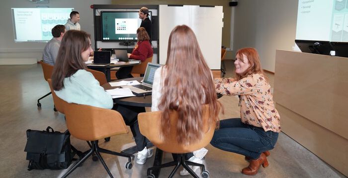 Bakerst i bildet ser vi en gruppe på fire studenter som som diskuterer. To av dem sitter rundt et bord, mens to stpr foran en digitavle. Foran i bildet ser vi to kvinnelige studenter som sitter rundt et bord og en kvinnelig underviser som sitter på huk og snakker med dem.