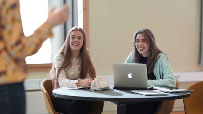 To studenter sitter ved et bord med notatbøker og en PC. Foran i bildet ser man en tredje person som snakker til studentene.