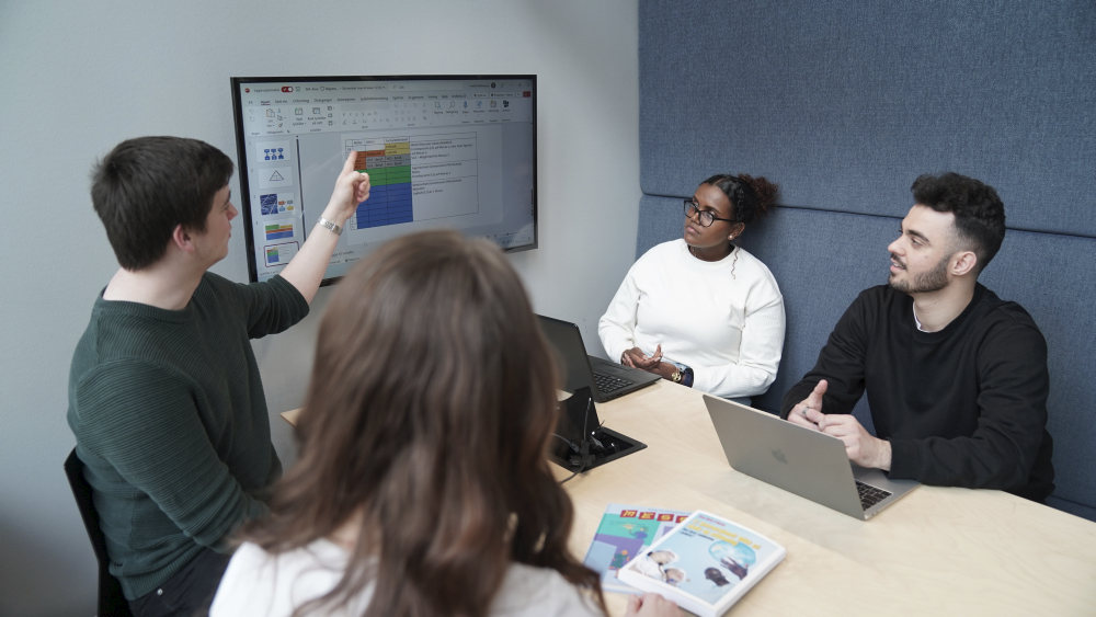 Fire studenter ved et gruppebord diskuterer PowerPoint-lysark på en stor skjerm