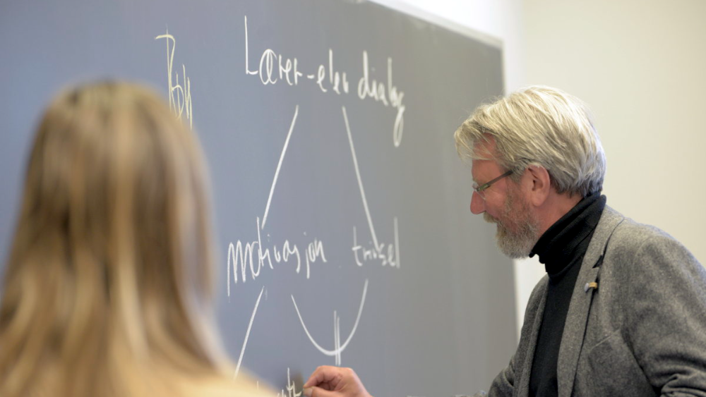 En gråhåret mannlig underviser som skriver på en tavle mens en annen person følger med på undervisningen.